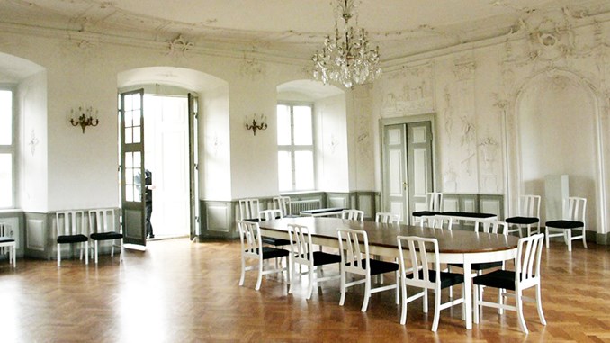 Havesalen på Augustenborg Slot med hvide vægge, stuk i loftet og stuk ned ad væggende. Store vinduespartier og en fransk dør ses til venstre i billedet. Der hænger en stor krystallysekrone i loftet.